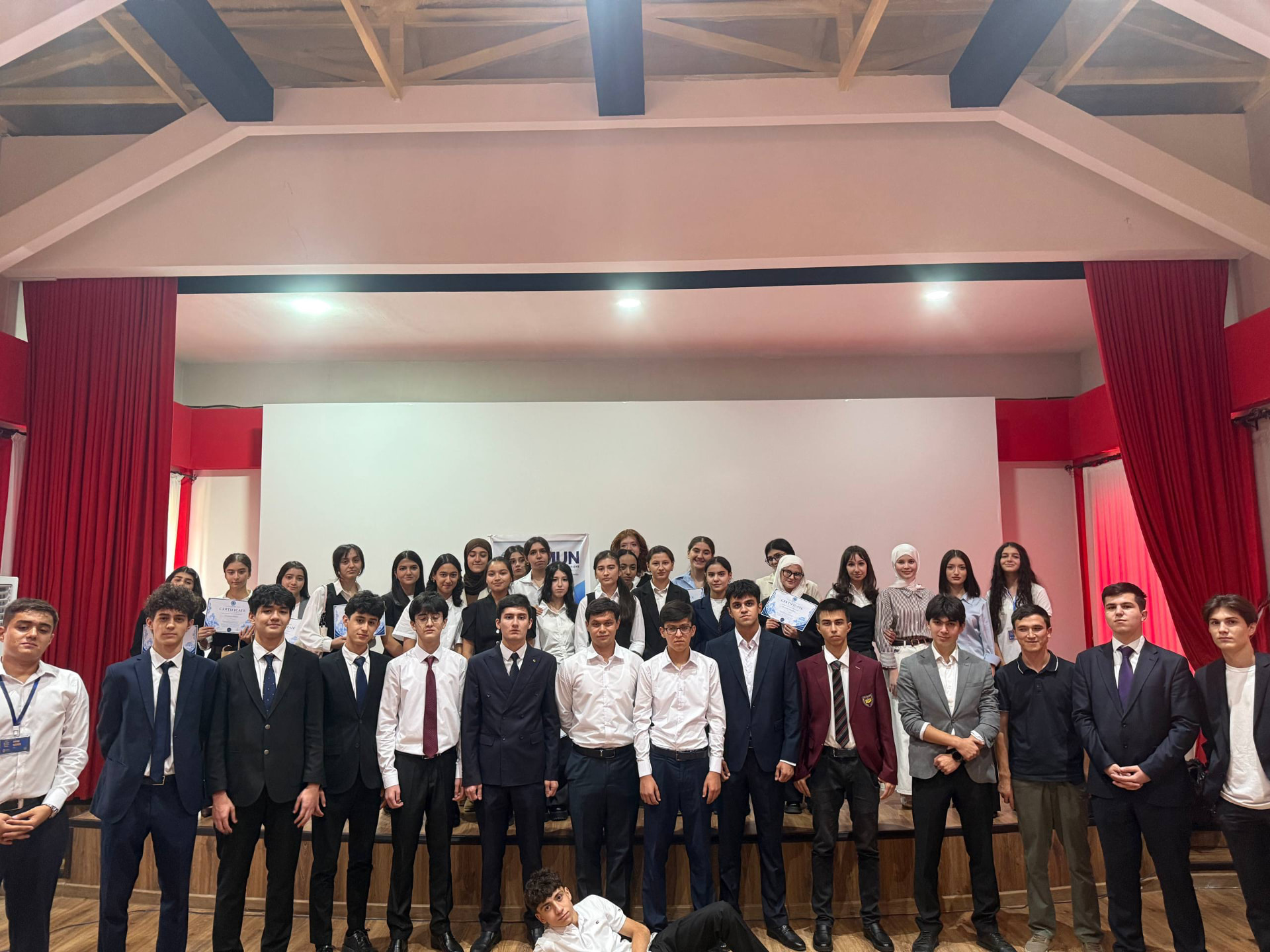 A large group of delegates in formal attire gathered on a stage, some holding certificates, in front of a DMUN banner.