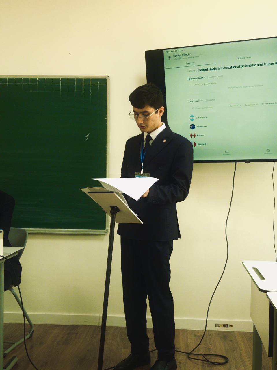 A delegate reading a speech from a lectern beside a screen showing a committee roster.